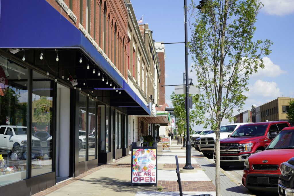Retail storefront street with multiple small shops, sidewalk signage, and parked cars in a downtown commercial area