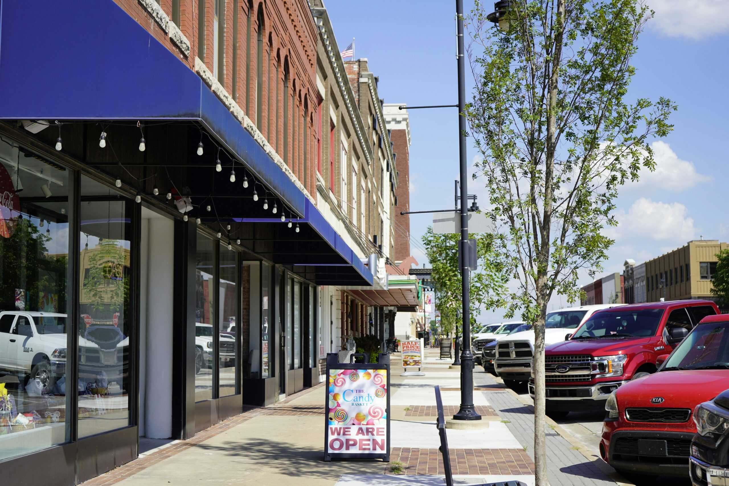 Retail storefront street with multiple small shops, sidewalk signage, and parked cars in a downtown commercial area
